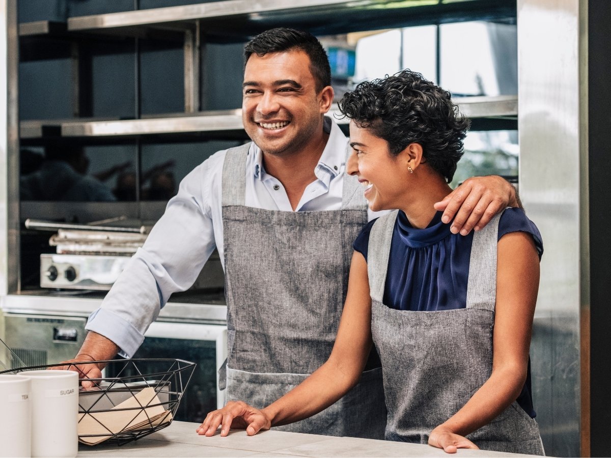Smiling man and woman wearing aprons standing together in a modern kitchen, appearing cheerful and engaged while preparing food.