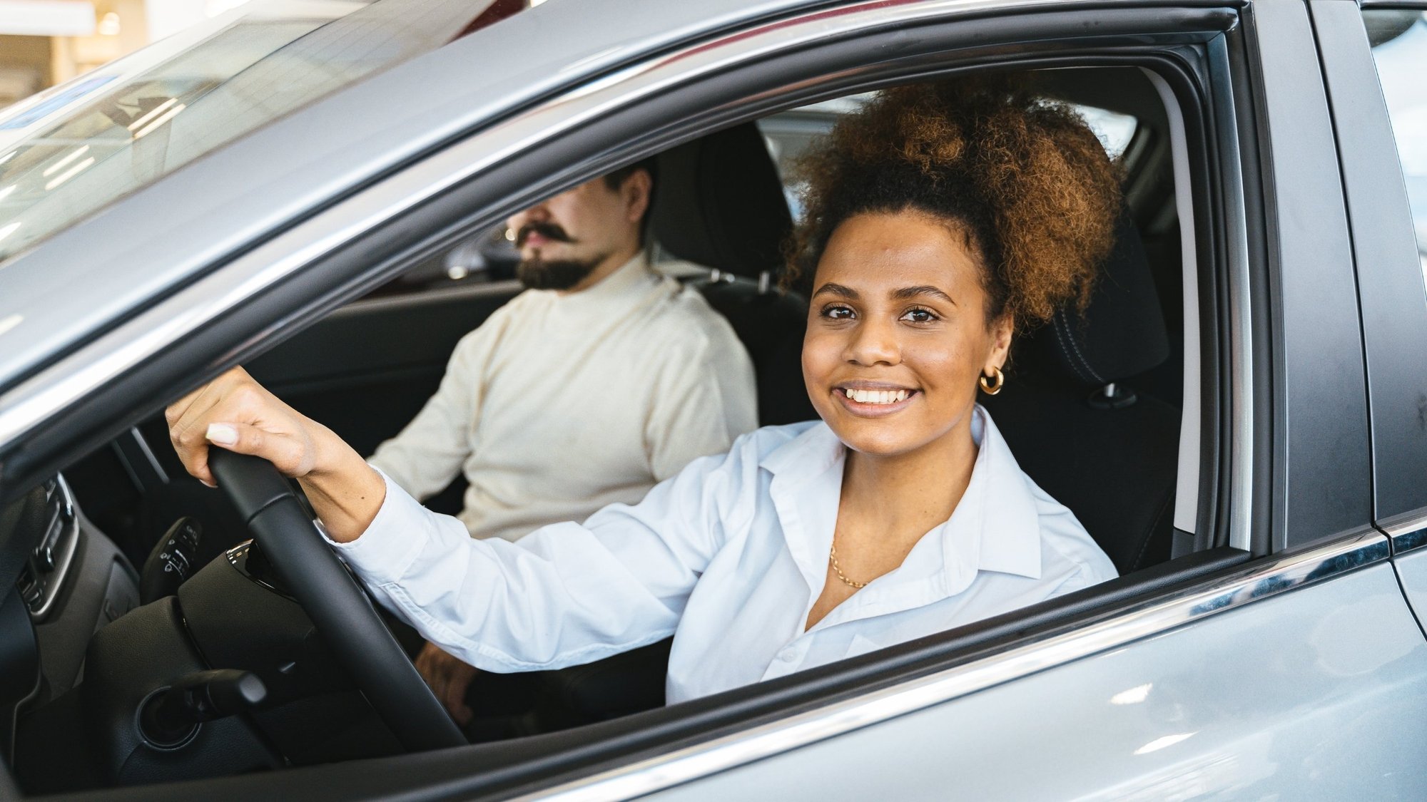 Smiling woman in the driver's seat of a car, holding the steering wheel and looking at the camera, with a man sitting in the passenger seat beside her.
