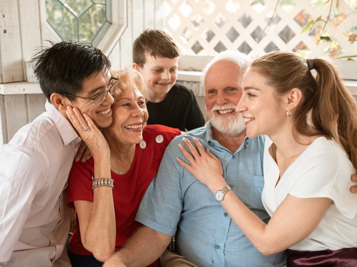 Multi-generational family sitting closely together on a porch, smiling and embracing each other warmly in a joyful, candid moment.