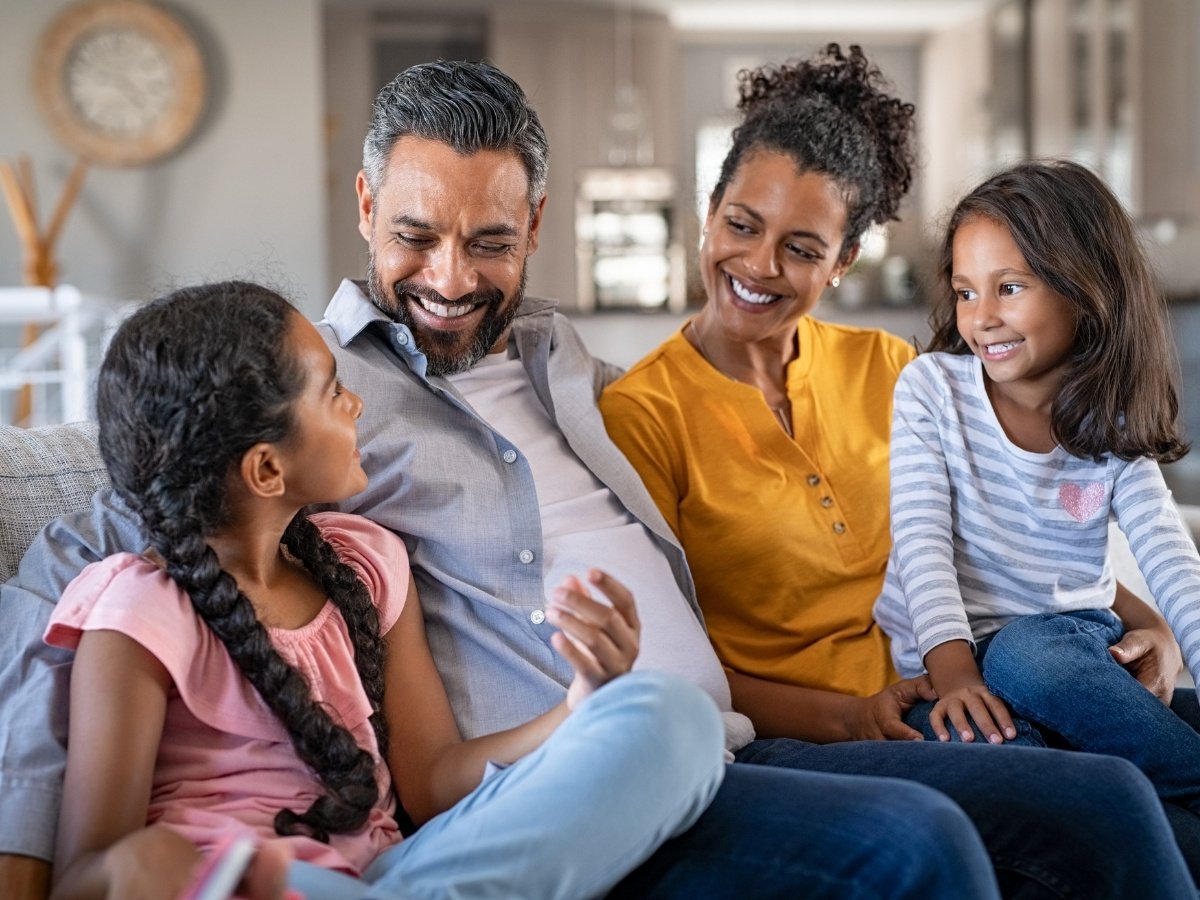 Happy family of four sitting together on a couch at home, smiling and talking with one another in a warm, relaxed setting.