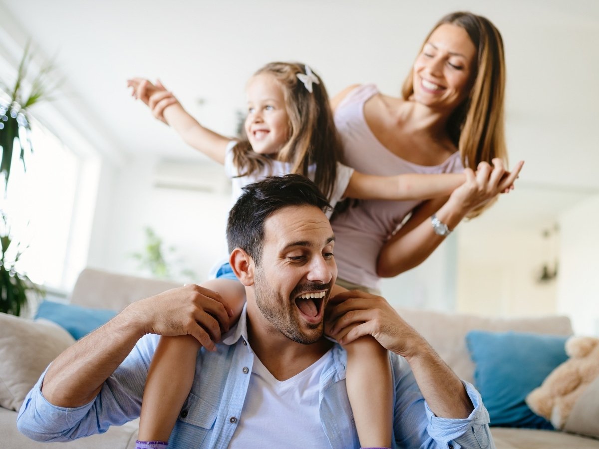 Happy family playing together at home, with a smiling father carrying his daughter on his shoulders while the mother laughs and joins in the fun.