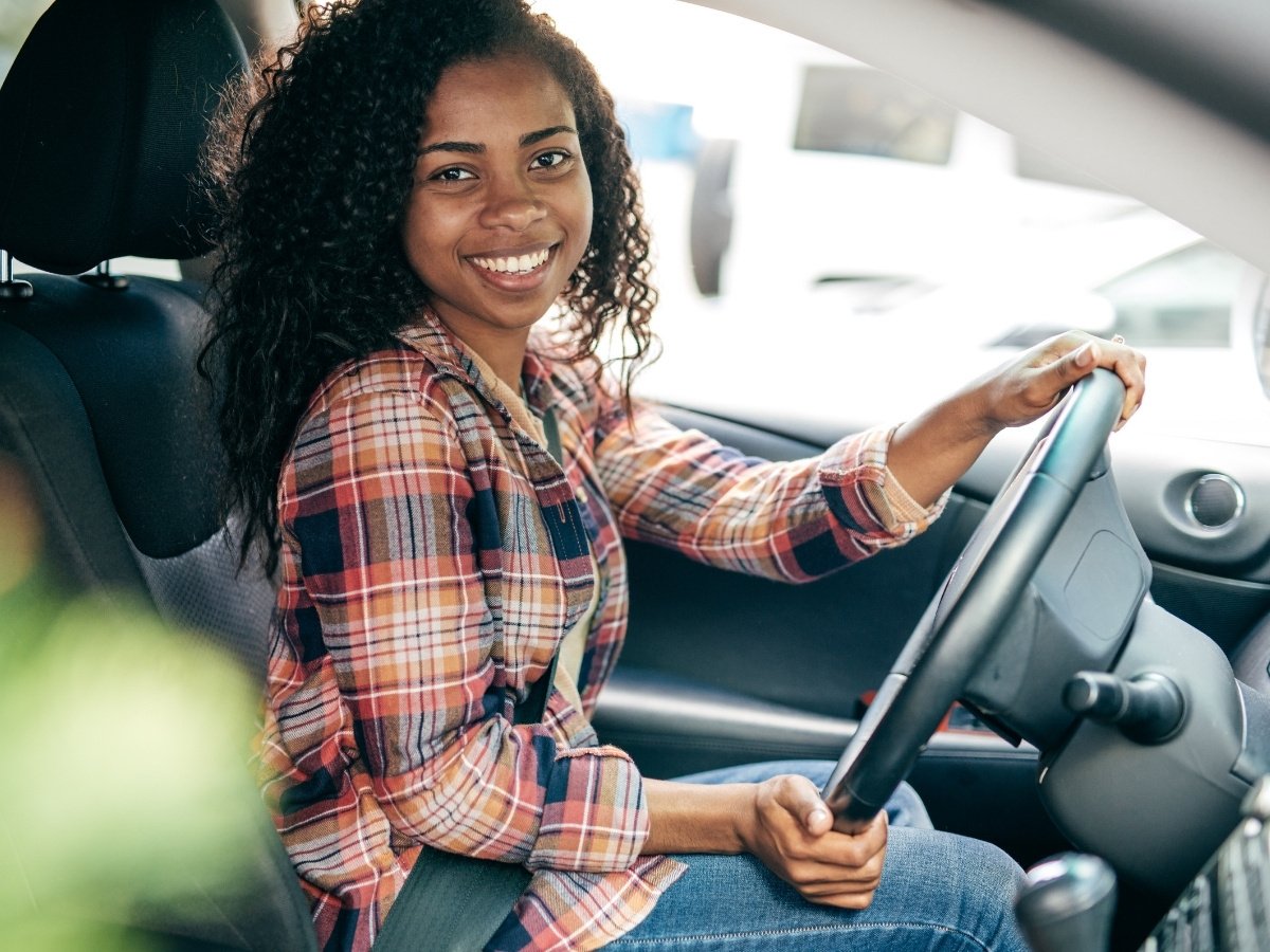 Smiling young woman sitting in the driver's seat of a car, wearing a plaid shirt and seatbelt, looking toward the camera with one hand on the steering wheel.