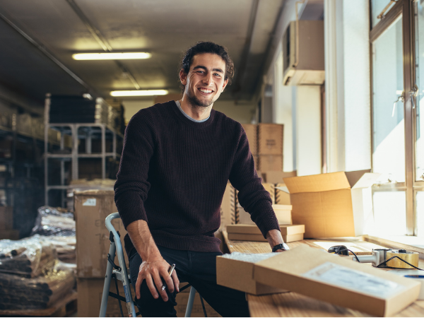 Smiling man sitting on a stool in a warehouse or packing area, surrounded by cardboard boxes and packaging materials, appearing relaxed and content while working on his business.
